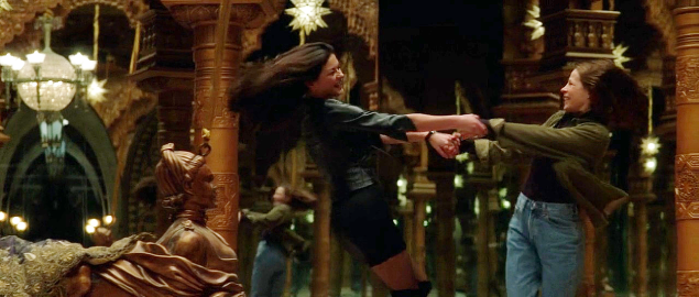 Two women, Nell and Theo, in an odd but ornate gold room with mirrored walls. They are holding hands and spinning around each other happily.